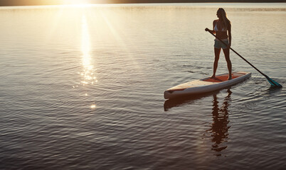 Lake, sunset and woman on paddle board for relax, water sports and vacation on river for tourism. Sunlight, summer and holiday in Germany for female person, swimsuit and travel fitness for adventure