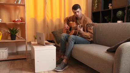 Adult man guitarist sitting in the room playing acoustic guitar watching lesson on laptop learning online. Hobby and leisure time concept.