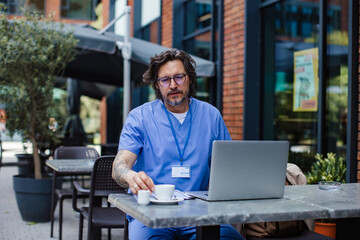 Obraz premium Doctor enjoying cup of coffee in coffee shop, working on laptop. Telemedicine consultation online. Mental health and life balance for a healthcare worker.