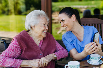 Female caregiver spending time with elderly woman, drinking coffee and talking. Nurse and senior...