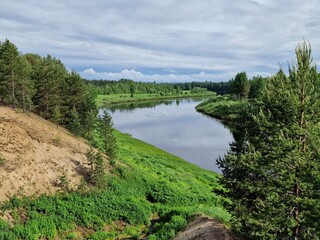A Winding River Through A Lush Forest Landscape on a Cloudy Summer Day. The river is calm and reflective, reflecting the green trees and the cloudy sky. The forest is dense and green, with tall trees 
