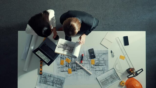 Top view of skilled project manager holding project plan and explain house design to architect engineer at meeting table with equipment, ruler, safety helmet and laptop scatter around. Alimentation.