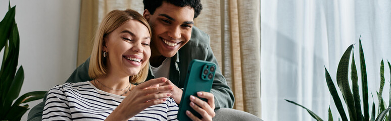 A young couple shares a laugh while looking at a smartphone in their modern apartment.