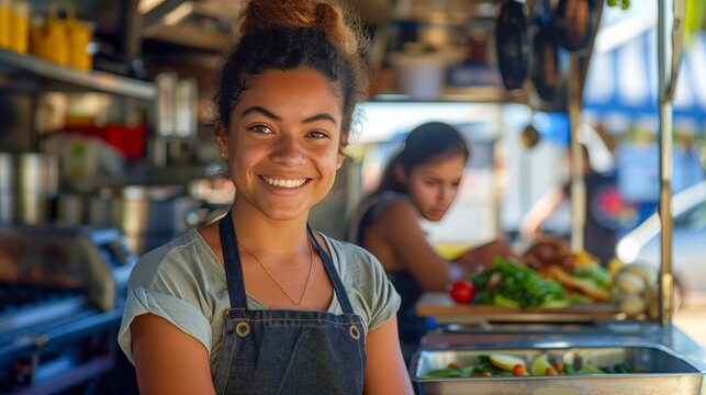 Smiling young woman cooking in a food truck, flanked by teammates preparing a salad and managing cargo.