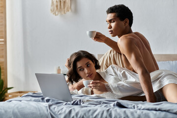 A young couple enjoys a relaxing morning together in bed, sipping coffee and using a laptop.