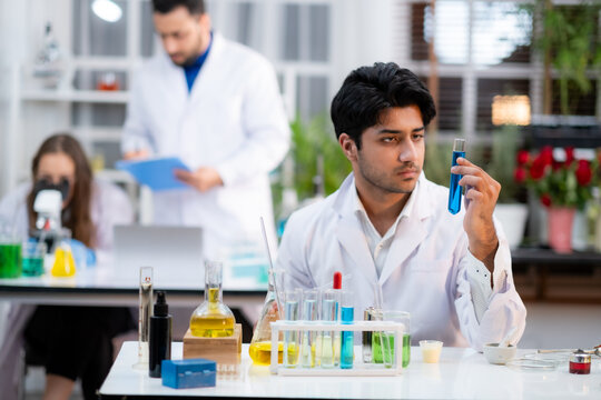 Focus at Young Scienctist man holding experiment tube with blue liquid looking at it with intense expression face.  With blur his co-workers working behide.