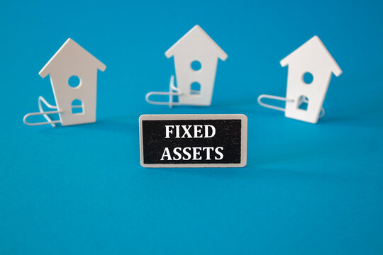 Fixed assets symbol. Wooden houses sits next to a wooden black board with the word fixed assets. Beautiful blue background. Business and fixed assets concept.