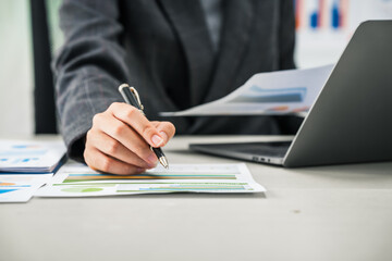 A businesswoman works diligently at her desk, reviewing financial documents and analyzing...