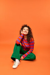 A young woman with curly hair sits on an orange background, wearing a striped sweater and green pants, smiling and looking up.