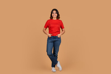 A young middle eastern woman stands in front of a tan background wearing a red t-shirt and blue...