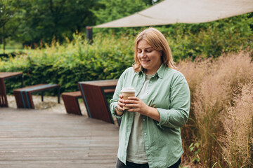 Happy redhead 30s Woman holding brown paper cup of coffee on public park background, Coffee to go on the city street. Food, rest, Take away concept.