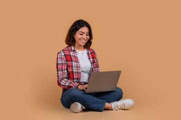 A young middle eastern woman sits on the floor with her legs crossed, smiling as she looks down at a laptop in her lap. The background is a light brown wall.