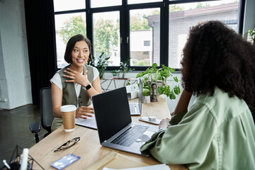 Two women sit at a table in an office, working together on a project.