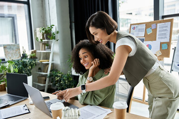 Two women work together on a laptop in a modern office.