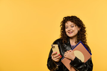 A young woman with curly hair, wearing a black leather jacket, smiles as she looks at her phone while holding a notebook.