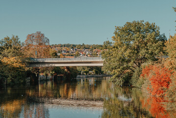 Beautiful small river with clean and clear water front of colorful autumn trees and small old town on the hill agaist nice blue and clouds sky during autumn in Europe