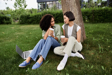 Two women sitting on the grass in a park, working on a laptop.