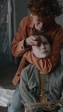 Vertical shot of young guy drawing cross with smouldering palo santo stick on forehead of devil possessed girl tied up to chair with rope during exorcism rites in derelict mansion
