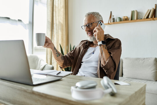 A mature gay man with grey hair and tattoos smiles while working remotely from home on his laptop.