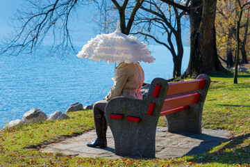 Woman with a Parasol Sitting on a Bench on the Waterfront to Lake Maggiore in a Sunny Day in Locarno, Ticino, Switzerland.