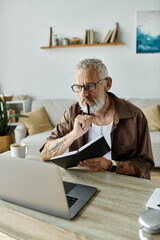 A mature gay man with tattoos and gray hair sits at a desk in his home office, working remotely while holding a notebook and pen.