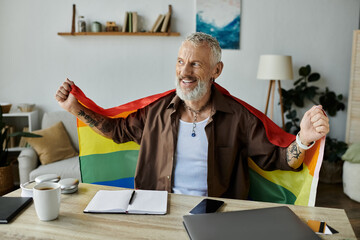 A mature gay man with tattoos and grey hair proudly holds a rainbow pride flag in his home.