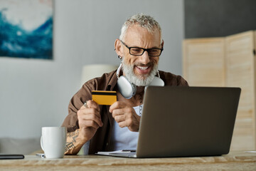 A mature gay man with tattoos and grey hair smiles as he shops online with a credit card, working remotely from home.