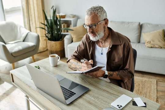 A mature gay man with tattoos and grey hair works on his laptop from home.