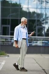 A mature man with a grey beard and tattoos walks down a city street, holding a laptop and checking his phone.