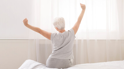 Senior woman sitting on her bed in morning, stretching with arms raised, back view, free space