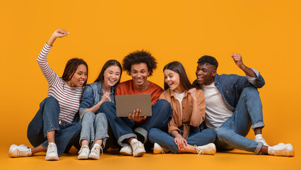 A group of five diverse friends sit on a yellow background, looking at a laptop and celebrating....