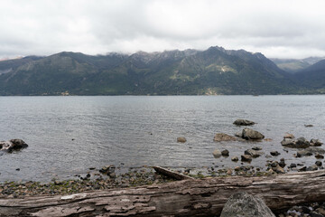 A large body of water with a rocky shoreline and a large mountain range in the background. The water is calm and the sky is cloudy