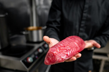 Close-up of a person’s hand presenting a raw, red piece of tuna in a kitchen setting, with a blurred stove in the background
