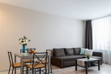 modern design of living room in apartment. Near window is big gray sofa with pillows, coffee table and plants. dining table and chairs near sofa, flowers in a vase, glasses and fruits on table