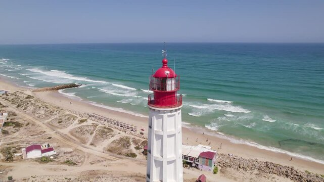 Drone orbits white lighthouse with red top as it overlooks sandy beach with strong ocean waves crashing on shore