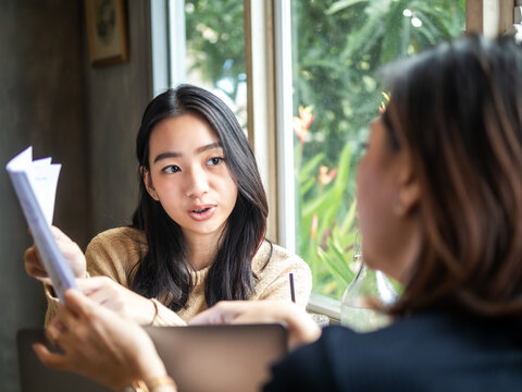 Asian woman in a café discussing and analyzing a report with a colleague. She looks concerned while holding a document with charts. The setting is modern and cozy, fostering a collaborative atmosphere