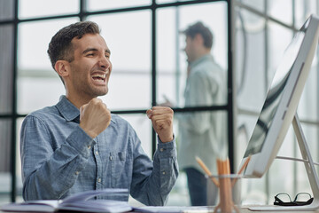 Happy businessman rejoicing success at workplace in office, looking at laptop screen with euphoric expression.