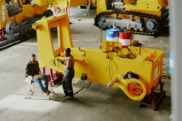 High angle view of two biracial coworkers sitting and standing by part of bulldozer machine using laptop and digital tablet