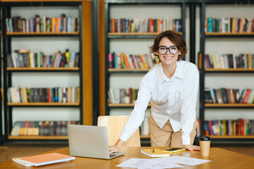 Young happy successful employee IT business woman she wearing glasses white shirt casual clothes use laptop pc computer stand near table work at office desk with pc laptop. Achievement career concept.