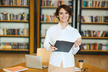 Young fun successful employee business woman wear white shirt casual clothes hold clipboard with paper account documents stand near table work at office desk with pc laptop Achievement career concept