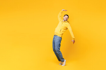 Full body side view young smiling happy man wearing hoody casual clothes stand on toes leaning back with outstretched hands dance isolated on plain yellow orange background studio. Lifestyle concept.