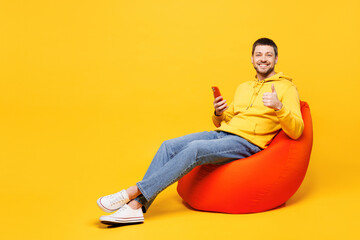 Full body young smiling happy man he wears hoody casual clothes sit in bag chair hold use mobile cell phone show thumb up isolated on plain yellow orange background studio portrait. Lifestyle concept.