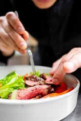 Close-up of a person’s hands cooking a salad with slices of medium-rare steak, fresh greens, and tomatoes in a white bowl.