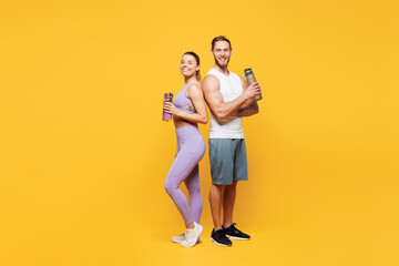Naklejka na ściany i meble Full body side view young strong fitness trainer sporty two man woman wear blue clothes spend time in home gym hold bottle drink water isolated on plain yellow background. Workout sport fit concept.