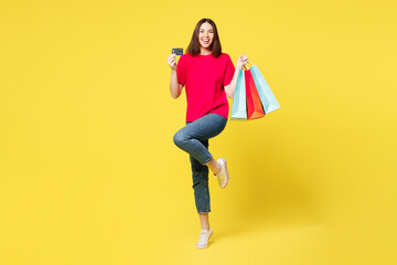 Full body young woman wears pink casual clothes hold in hand paper package bags after shopping, show mock up of credit bank card isolated on plain yellow background. Black Friday sale buy day concept.