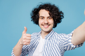 Close up young fun man wear shirt white t-shirt casual clothes doing selfie shot pov on mobile cell phone show thumb up isolated on plain pastel light blue cyan background studio. Lifestyle concept.