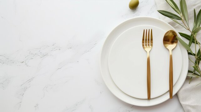 A white marble tabletop with a white plate, gold fork, gold knife, and gold spoon. An olive branch with green leaves extends from the left side of the frame