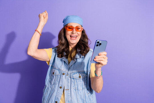 Photo portrait of attractive senior woman hold device winning dressed stylish jeans hipster clothes isolated on purple color background