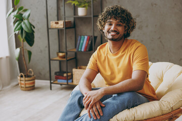 Young smiling happy cheerful fun Indian man he wearing orange casual clothes look camera sits in armchair stay at home hotel flat rest relax spend free spare time in living room indoor Lounge concept