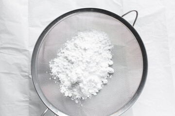 Overhead view of icing sugar in a silver sieve, sifting powdered sugar on a white table, top view of powdered sugar being sieved on a marble countertop